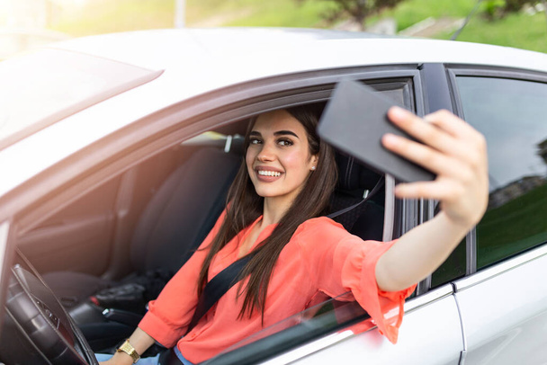 Smiling young woman taking selfie picture with smart phone camera outdoors in car. Holidays and tourism concept - smiling teenage girl taking selfie picture with smartphone camera outdoors in car - Fotó, kép
