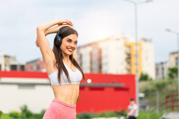 Athletic young woman with head phones smiling and streaching. Young sporty woman training. Woman Doing Workout Exercises On Street . Fit girl streaching before active fitness training - Photo, Image