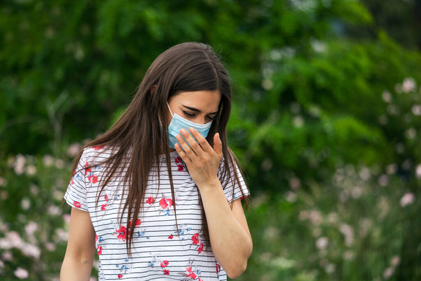 Portrait of young woman wearing a protective mask sneezes on green nature background with flowers in protective medical mask, coronavirus pandemic - Photo, Image