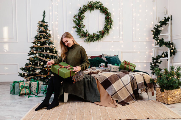 Μια νεαρή γυναίκα ανοίγει χριστουγεννιάτικα δώρα σε μια ατμόσφαιρα Πρωτοχρονιάς και χαίρεται. Η γυναίκα είναι σε ένα φωτεινό, άνετο υπνοδωμάτιο, όμορφα διακοσμημένο για τα Χριστούγεννα. - Φωτογραφία, εικόνα