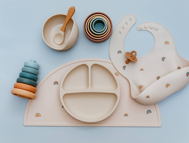 Pastel silicone collection of tableware, bibs, accessories, Montessori toys for children on blue background. Baby accessories, tableware concept. - 写真・画像