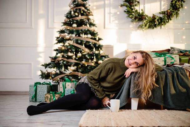 Pretty young woman with blond hair sits near a Christmas tree with coffee. New Year is coming soon. Christmas atmosphere in a cozy home - Foto, afbeelding