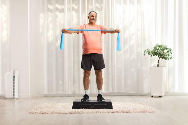 Mature man in sportswear exercising with a stretch strap on a step aerobic platform inside a room - Φωτογραφία, εικόνα