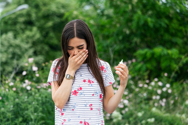 Young pretty woman sneezing in front of blooming tree. Spring allergy concept. Woman applies nasal spray. Pollen allergy symptoms - Photo, image