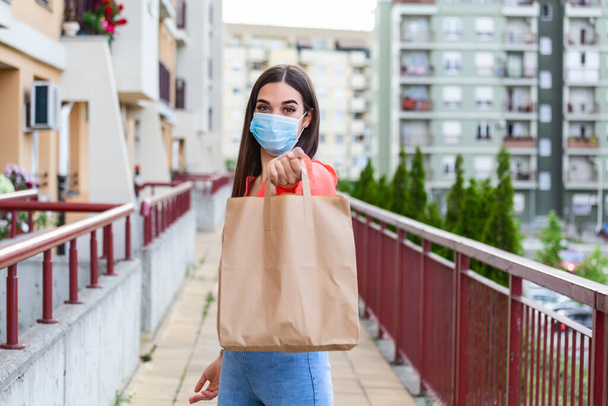 Young woman volunteer with a medical mask is holding a paper bag with food and vegetables. Donation, helping people in quarantine, coronavirus. Delivery shopping during Covid 19 outbreak. - Foto, Imagem