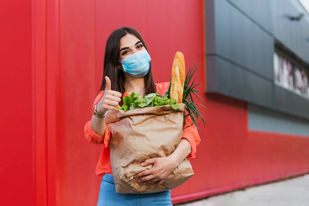 Pretty girl with medical mask holding bag with groceries and looking at camera with thumb up. Woman holding heavy bag with groceries. Groceries shopping during Covid 19, coronavirus pandemic - Foto, Imagen