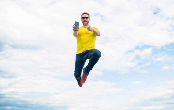 man jumping and presenting smartphone on sky background. - 写真・画像