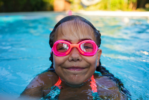 Portrait of girl with two braids in swimming suit and goggles smiles and enjoy training swim at pool in summer camp. Refreshing at heat weather, active vacation and healthy lifestyle, happy summertime - Foto, afbeelding