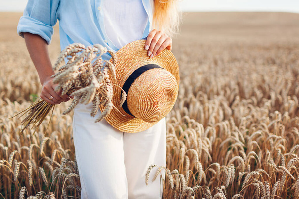 Close up of summer female accessory and clothes. Woman holding straw hat wearing linen shirt in wheat field at sunset. Natural hypoallergenic clothing. Rustic boho style - Foto, Bild
