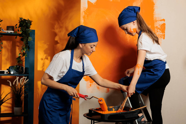 Woman and girl renovating house with paint, using orange color to decorate wall surface in apartment. Young child on ladder painting with paintbrush and adult with rolling brush, having fun. - Foto, imagen