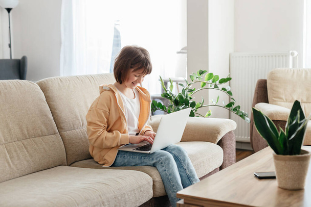 Ensino em casa para crianças. Uma menina se senta em um sofá com um laptop no colo e assiste webinars ou bate-papos com amigos on-line. Crianças usam tecnologia - Foto, Imagem