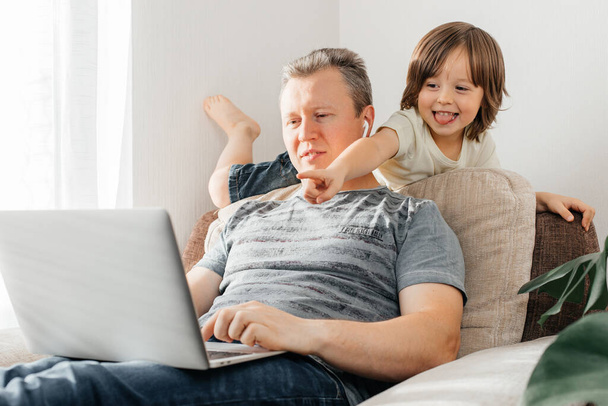 Be a freelance parent. Dad sits on an armchair and works at a laptop, a little son sits on dad and has fun. Remote work from home. - Фото, зображення