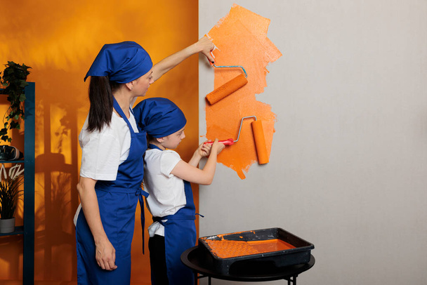 Young people painting walls with orange color paint and roller brush, doing housework renovation together. Mother with little kid using paintbrush and redecorating tools in apartment. - Φωτογραφία, εικόνα