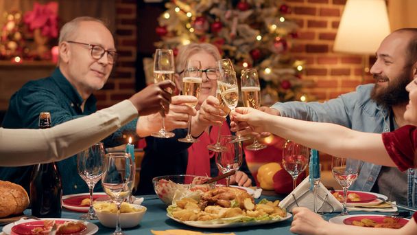 Diversos miembros de la familia festivos disfrutan de la cena de Navidad en casa mientras beben champán. Gente multicultural alegre celebrando la fiesta de invierno con comida casera y vino espumoso. - Foto, Imagen