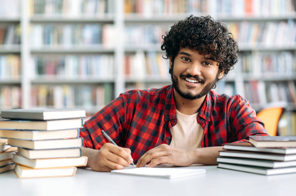 Back to school. Happy clever Indian or Arabian male student, in stylish wear, sits at the table with books in the university library, takes notes in a notebook, looking at the camera, smiling friendly - Fotografie, Obrázek