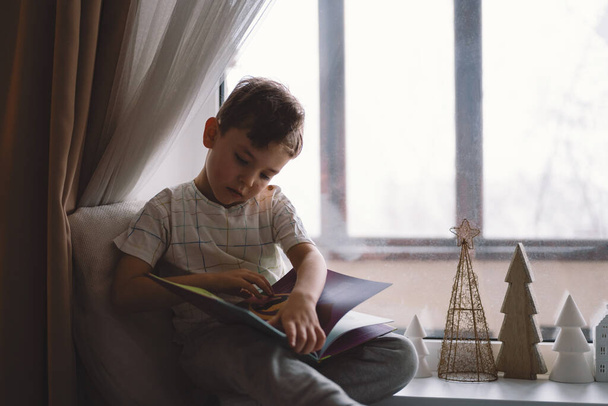 Cute boy is reading a book sitting on the windowsill near the window. Cozy home with decorative Christmas trees. Sweet home. Winter holidays lifestyle. - Foto, imagen