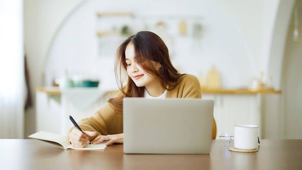 Μια νεαρή Ασιάτισσα κάθεται στο τραπέζι και κρατάει σημειώσεις στο σημειωματάριο. Δουλεύοντας στο τραπέζι με το laptop του υπολογιστή, έγγραφο γραφειοκρατία και φλιτζάνι καφέ. Μαθητεία online - Φωτογραφία, εικόνα