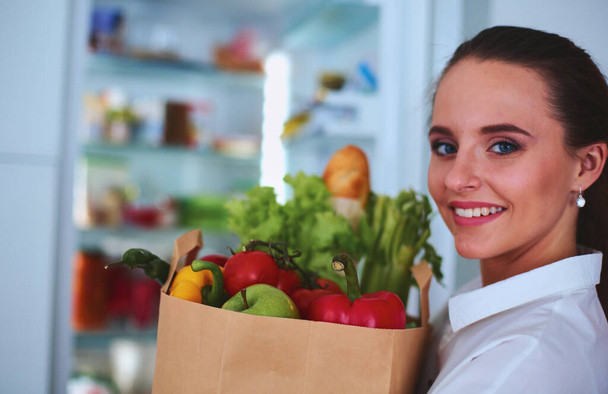 Mladá žena drží nákupní tašku s potravinami se zeleninou.Stojící v kuchyni. - Fotografie, Obrázek
