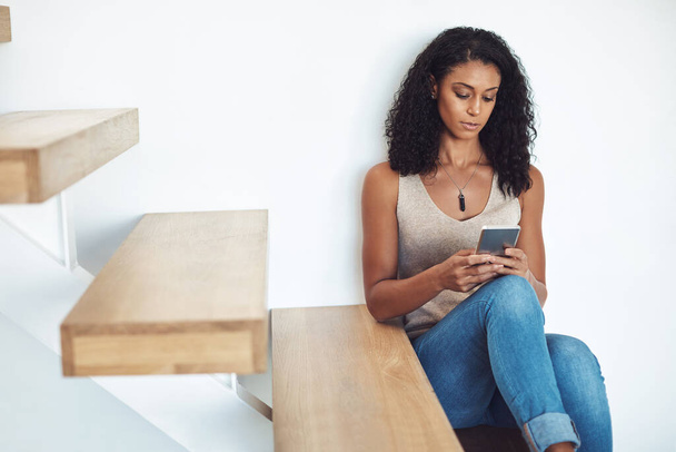 Woman relaxing, looking and typing on phone sitting on wooden staircase. Attractive, casual and confident African American female reading on mobile. Taking a break and catching up on social media. - Photo, image