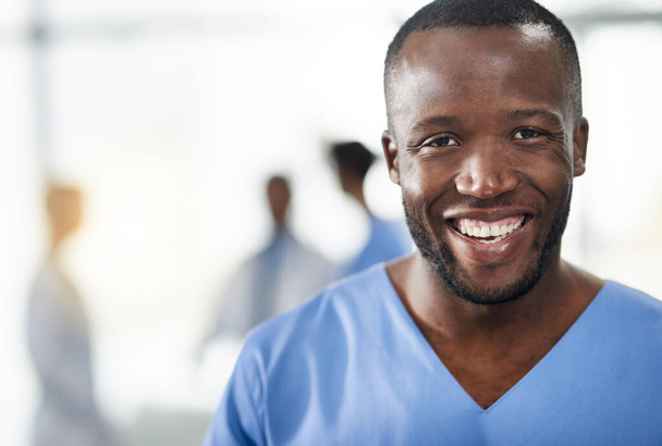 Happy, smiling and professional doctor in a hospital closeup portrait with blurred background. Confident black male medical healthcare worker with colleagues in the back. An African American surgeon - Фото, зображення