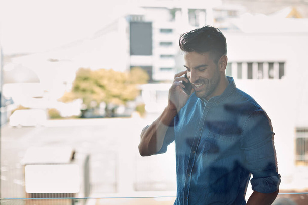 Профессиональный бизнесмен улыбается по телефону, в современном офисе. Очаровательный, уверенный в себе сотрудник, держит мобильный телефон и разговаривает во время мобильного разговора. Работник, стоящий на улице на рабочем месте. - Фото, изображение