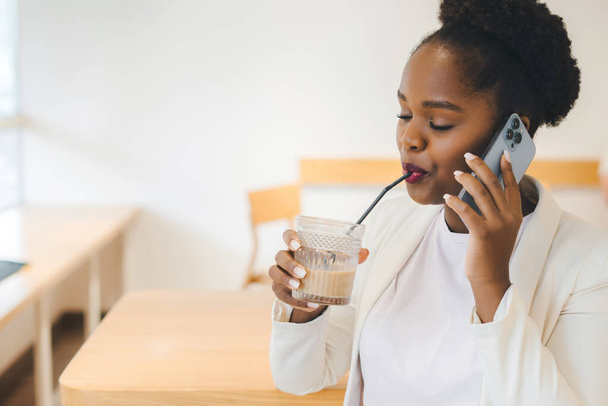 Positive afro woman having conversation on mobile phone enjoying tasty fresh drink at coffee shop. Beauty portrait. Beauty concept. Attractive beautiful - Zdjęcie, obraz
