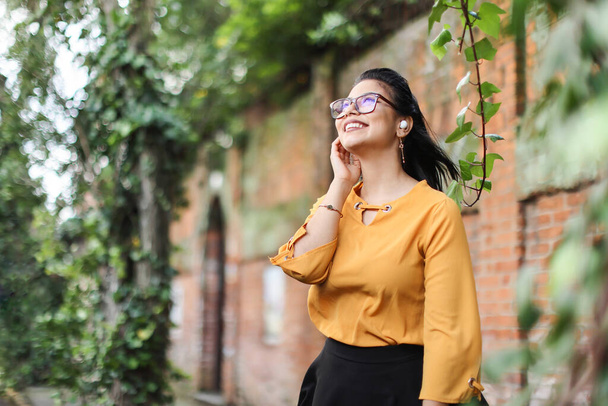Young Latin American woman wearing eyeglasses and earbuds looking up smiling with leaves copy space. High quality photo - Photo, image