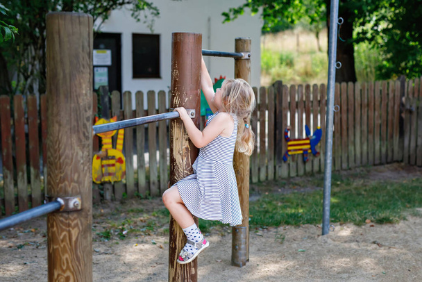 Little preschool girl climbing and hanging on outdoor horizontal bar. Cheerful girl athlete on gymnastic stick on sports ground. Happy child and summer activities on playground - Foto, immagini
