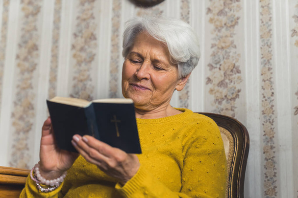 Positive elderly retired female reading a book sitting on a chair. Peaceful retirement. High quality photo - Foto, afbeelding
