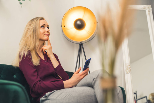 Blonde white woman wearing maroon shirt in living room putting pinger to chin thinking and looking up holding smartphone. Indoor shot. High quality photo - Fotoğraf, Görsel