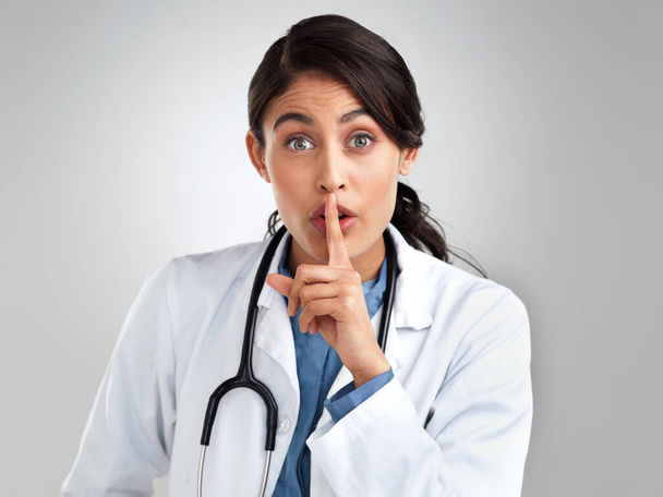 Dont ask, dont tell. Studio portrait of a young doctor with her finger on her lips against a grey background - Foto, Imagen
