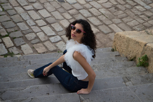 schöne junge Frau mit dunklen, lockigen Haaren, die auf einer Treppe in einem Park sitzt. Die Frau trägt eine Sonnenbrille und macht unterschiedliche Mienen. Ausdruck und Lifestylekonzept - Foto, Bild