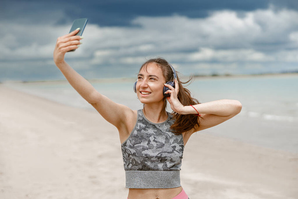 Woman in sportswear standing on beach and making selfie on background of ocean. High quality photo - Фото, изображение