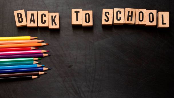 square wooden letters with the phrase "Back To School" written in English on a black chalkboard background and next to colored pencils - Zdjęcie, obraz