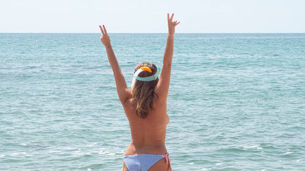 Young woman from the back in a swimsuit raising her arms making the victory symbol on the beach with blue water - Foto, Bild