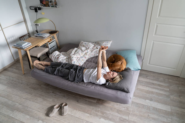 Lazy middle-aged woman lies on cozy bed near sleeping dog. Mature woman wearing pajama raises hands without desire of getting up. Unwashed lady wants to sleep on double bed upper view. Morning routine - Foto, Imagem