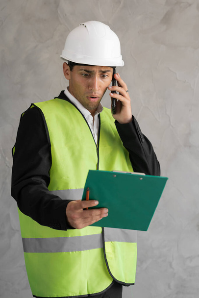 Angry muslim engineer or architect screaming while talking on the phone, builder holding clipboard and cursing into smartphone, builder isolated on gray background - Foto, Imagen
