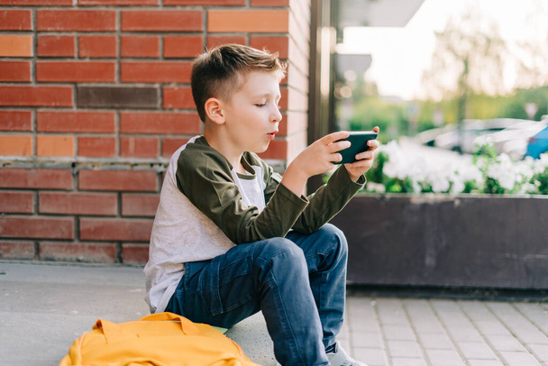 Back to school. Cute child with backpack, holding mobile phone, playing with cellphone. School boy pupil with bag. Elementary school student after classes. Kid sitting on stairs outdoors in the street - Фото, изображение