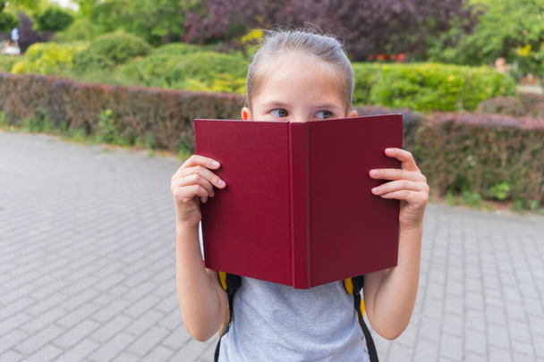 Smiling 6-8-year-old girl with long hair, with backpack and book, dressed in gray T-shirt, stands near the school, having made roof of house over her head from book. The concept of Back to School. - Photo, image