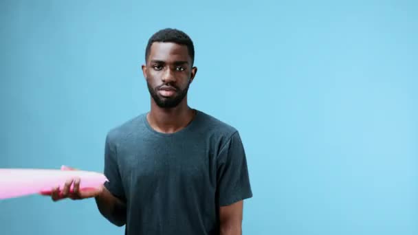 Hombre afroamericano juega con un bate inflable de color rosa en sus manos, se golpea en la cabeza, baila descuidadamente en una camiseta en azul. fondo en el estudio. Imágenes de alta calidad 4k - Metraje, vídeo