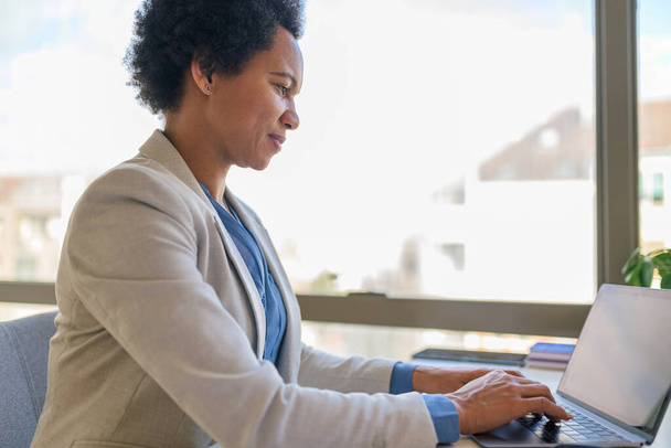 Confident female executive working on laptop. Entrepreneur with afro hair is wearing formals. She is sitting at desk in corporate workplace. - 写真・画像