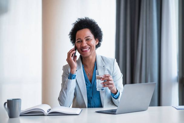 Portrait of smiling businesswoman talking on smart phone. Female executive wearing formals is having glass of water. She is working on laptop at desk in the office. - Photo, Image