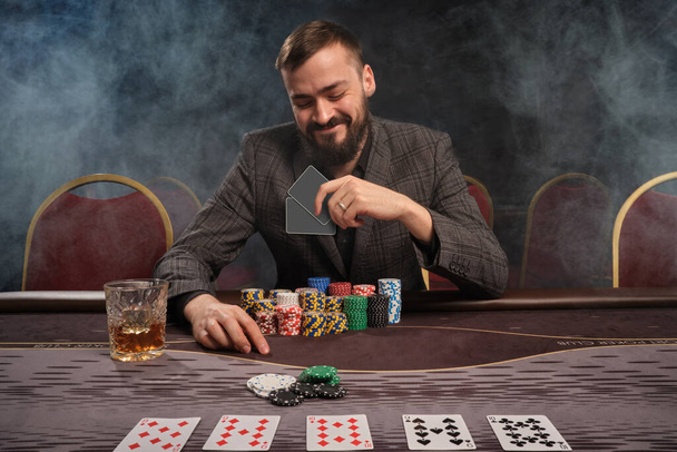 Bearded person in a gray suit is playing poker sitting at the table at casino in a smoke. He is holding two cards in his hand and smiling looking at it. Gambling addiction. Entertainment concept. - Photo, Image