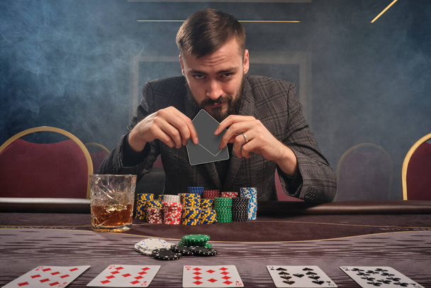 Bearded guy in a gray suit is playing poker sitting at the table at casino in a smoke. He is holding two empty cards in his hand and looking at the camera. Gambling addiction. Entertainment concept. - Photo, Image