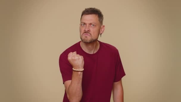 Aggressive angry handsome young man in red t-shirt trying to fight at camera, shaking fist, boxing with expression, punishment. Adult stylish male guy isolated alone on beige studio background indoors - Filmagem, Vídeo