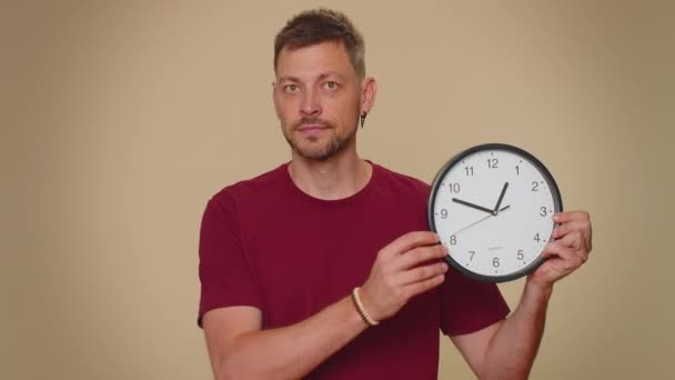 It is your time. Handsome young man in red t-shirt showing time on wall office clock, ok, thumb up, approve, pointing finger at camera. Adult stylish male guy on beige studio wall background indoor - Metraje, vídeo
