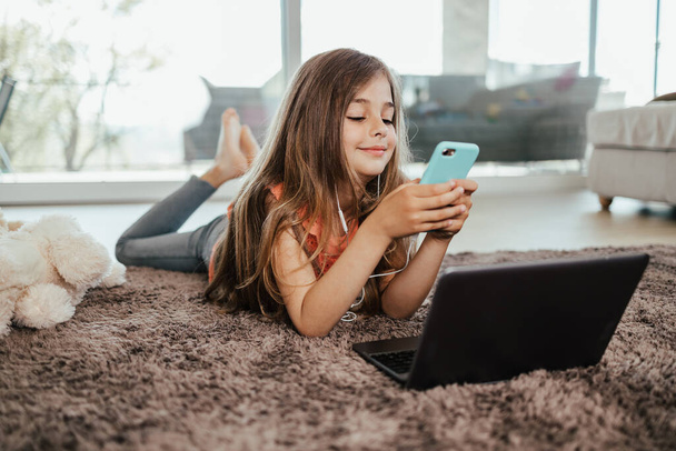 Beautiful girl lies on the floor of the living room of her house and uses a laptop and smart phone for learning and communication. She is happy and smiled. - Foto, imagen