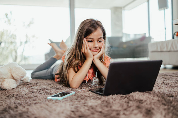 Beautiful girl lies on the floor of the living room of her house and uses a laptop and smart phone for learning and communication. She is happy and smiled. - Photo, image
