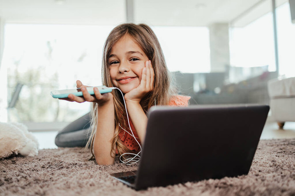 Beautiful girl lies on the floor of the living room of her house and uses a laptop and smart phone for learning and communication. She is happy and smiled. - Photo, Image