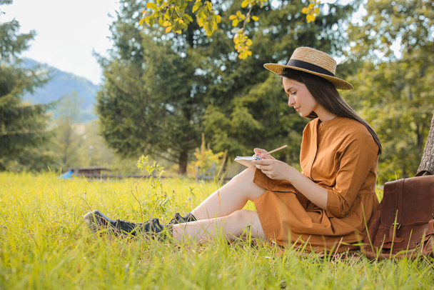 Beautiful young woman drawing with pencil in notepad outdoors on green grass - Photo, image